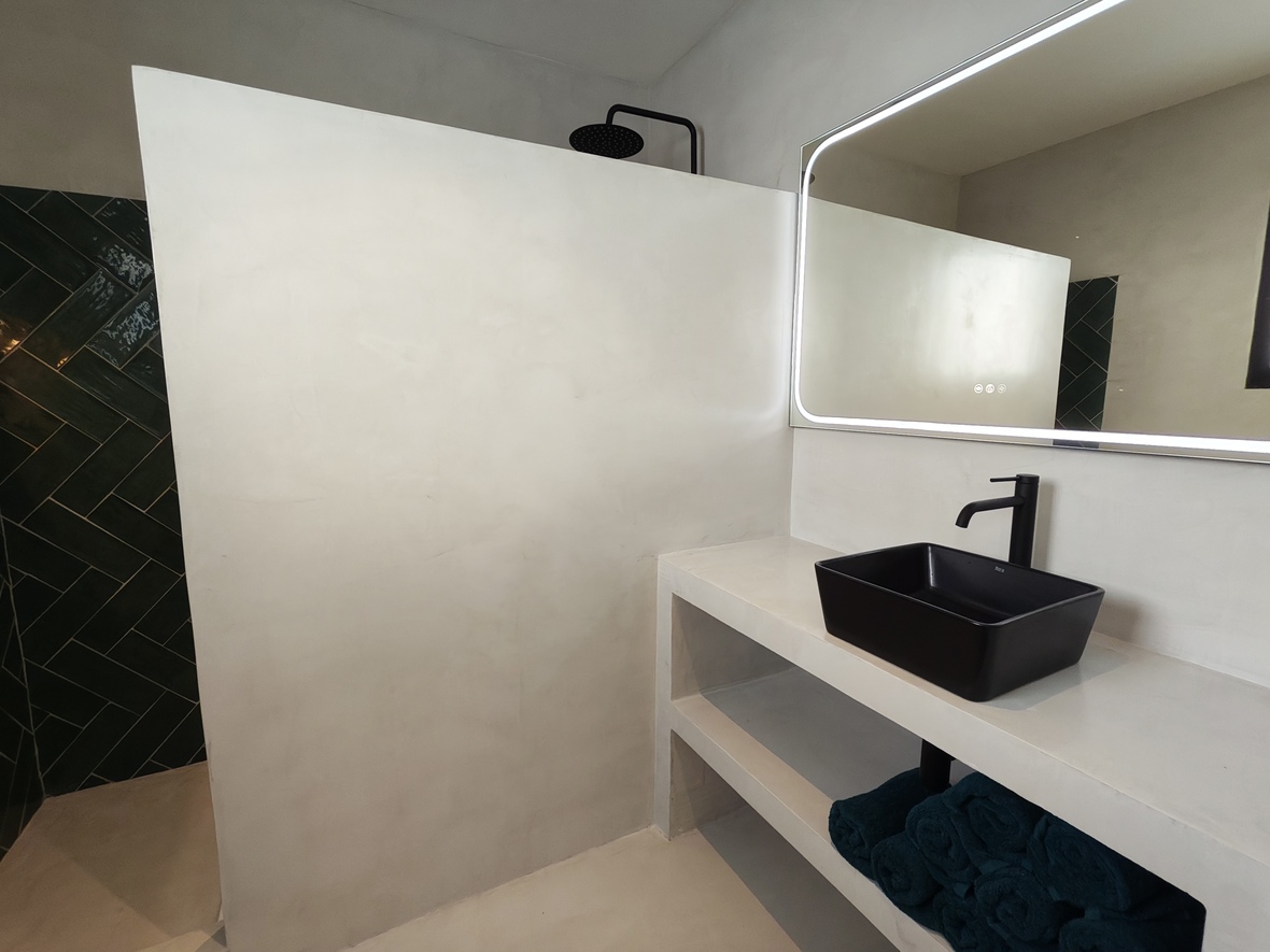 Modern bathroom featuring microcement finishes, a black vessel sink, and a backlit LED mirror. The walk-in shower includes dark teal herringbone tiles and matte black fixtures.