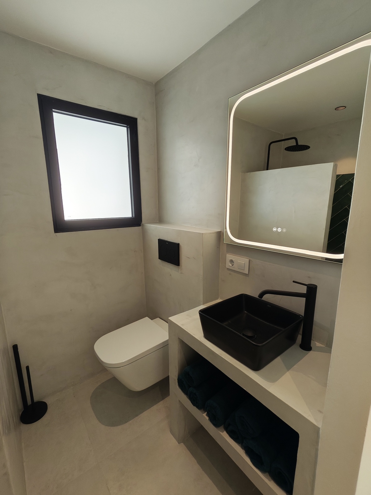 Modern bathroom featuring microcement walls, a black matte vessel sink, and a backlit LED mirror. Includes black fixtures and a wall-hung toilet.