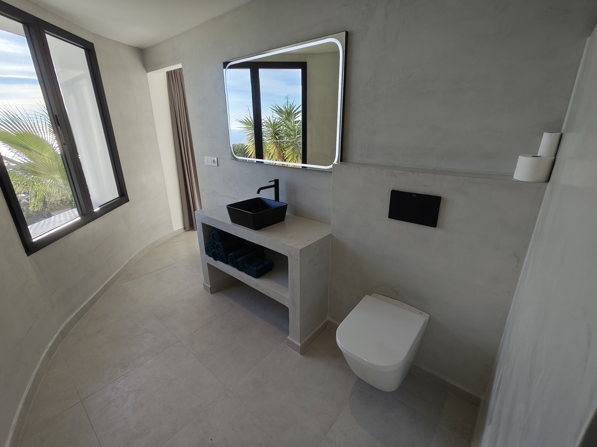 Modern bathroom featuring microcement walls, a black vessel sink on a stone vanity, and a backlit mirror. Large windows provide natural light and palm tree views.