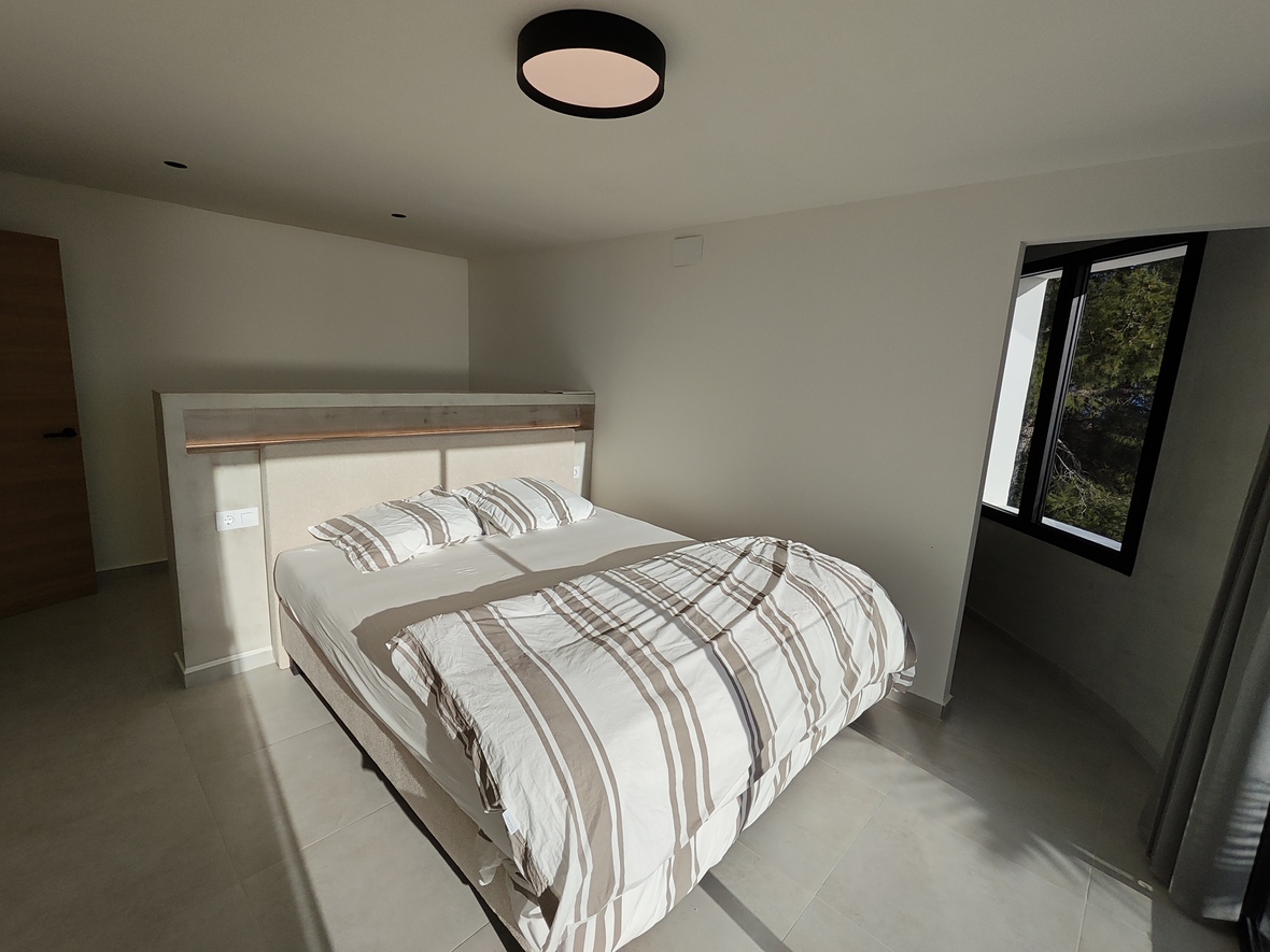 Modern bedroom featuring a custom partition headboard with integrated LED lighting, large format tile flooring, and a black-framed window overlooking greenery.