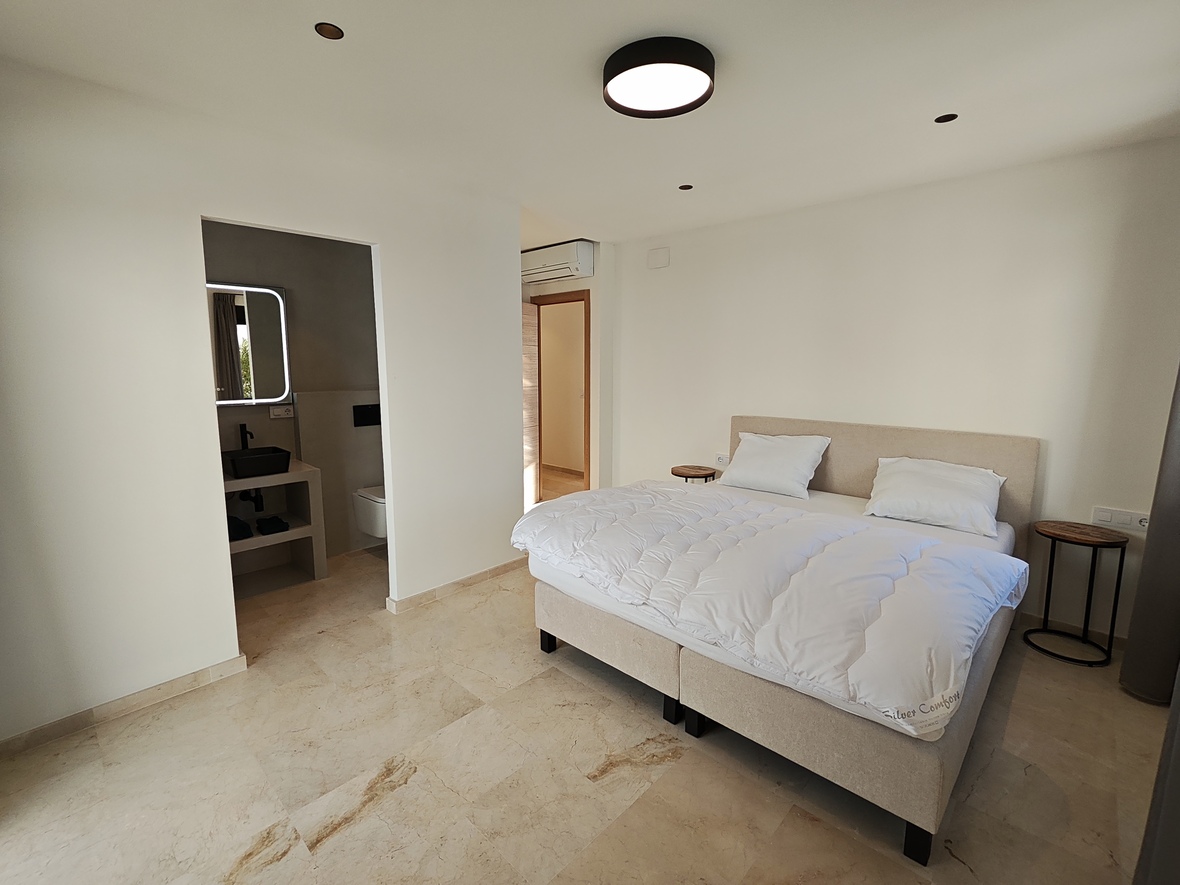 Modern bedroom featuring polished marble flooring, an upholstered double bed, and an integrated en-suite bathroom with a black vessel sink and backlit mirror.