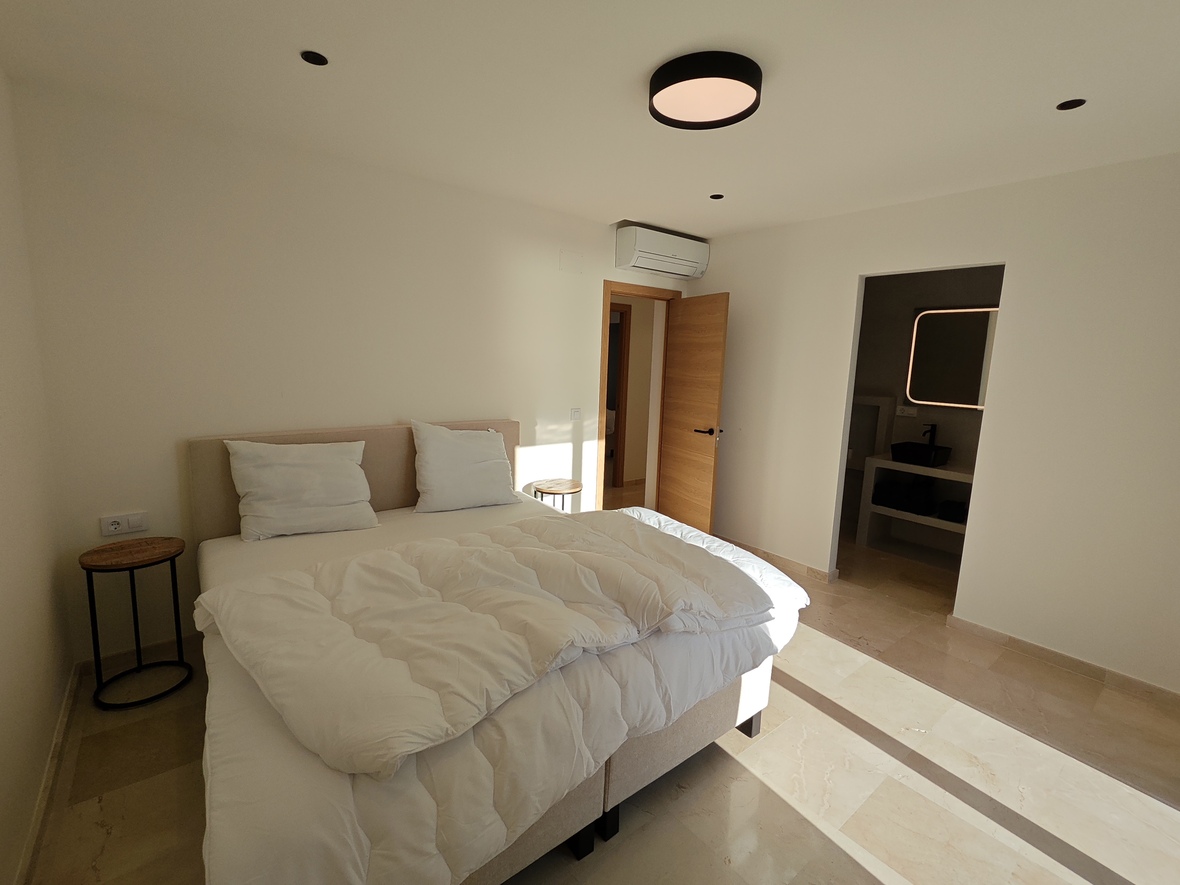 Modern bedroom featuring stone tile flooring, a beige upholstered headboard, and minimalist side tables. The space includes air conditioning and an en-suite bathroom with a backlit mirror.