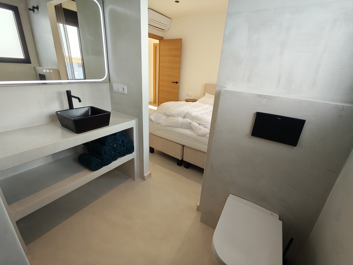 Modern ensuite bathroom featuring microcement finishes, a black vessel sink, and a backlit mirror, with a view into the adjacent bedroom.