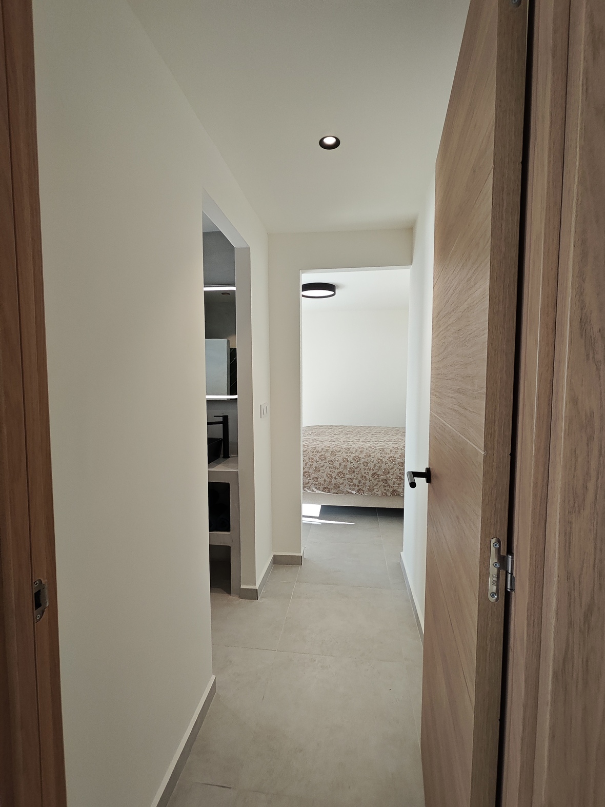 Modern hallway with light wood doors and grey tiled flooring, offering access to a minimalist bathroom and a bright bedroom with floral bedding.