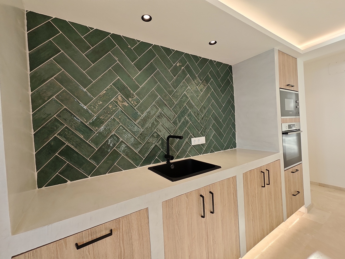 Modern kitchen featuring a green herringbone tile backsplash, black sink, and light wood cabinetry with integrated appliances and recessed lighting.