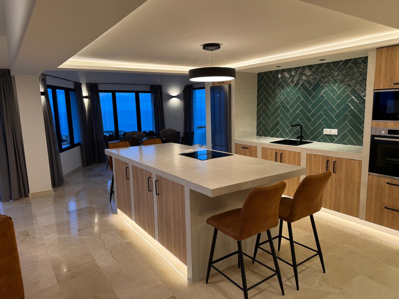 Modern kitchen featuring a microcement island, green herringbone tile backsplash, and panoramic sea views through large windows at dusk.