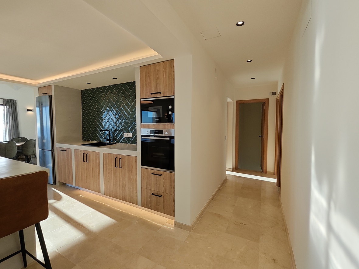 Modern kitchen featuring wood cabinetry, a dark green herringbone tile backsplash, and integrated appliances, adjacent to a bright hallway with stone flooring.