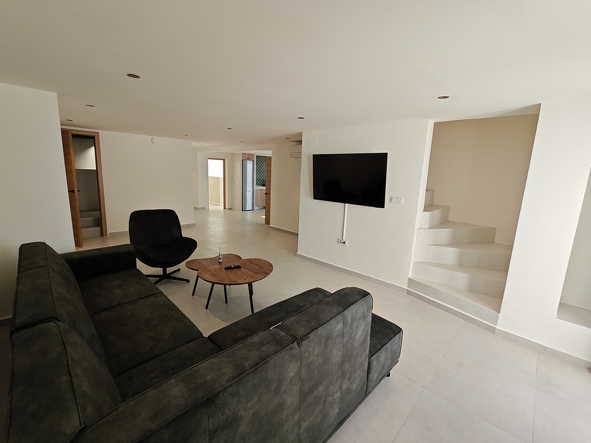 Modern living area featuring a dark sectional sofa, wooden coffee table, and a wall-mounted TV adjacent to a minimalist white staircase with recessed lighting.