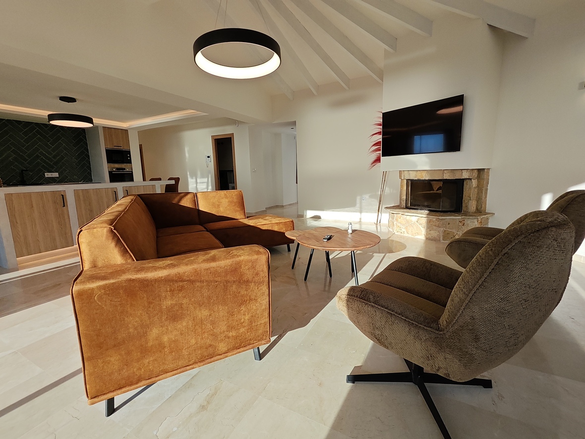 Modern living room featuring an ochre velvet sofa, stone-clad fireplace, and vaulted beamed ceilings. An open-plan kitchen with green herringbone backsplash is visible in the background.