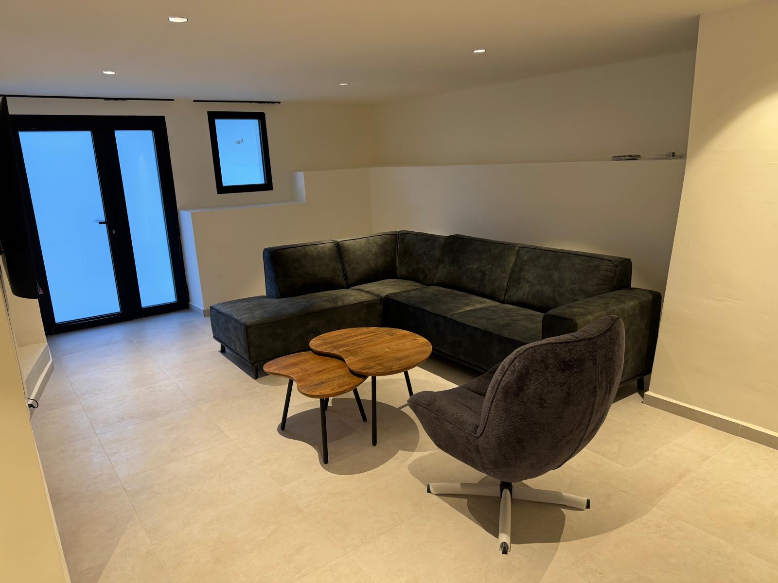 Modern lounge area featuring a dark grey sectional sofa, organic wooden coffee tables, and a swivel armchair on light tiled flooring with recessed lighting.