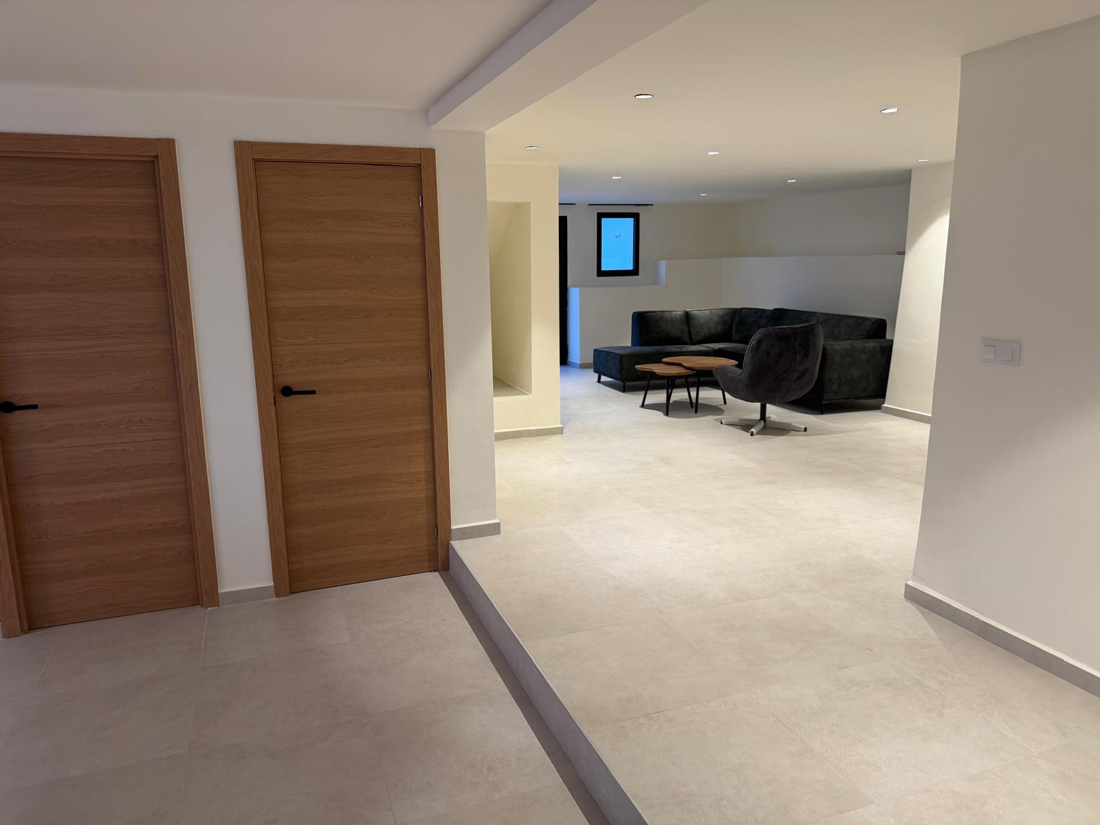 Modern lounge area featuring light grey tiled flooring, recessed lighting, and a black sectional sofa complemented by oak-toned interior doors.