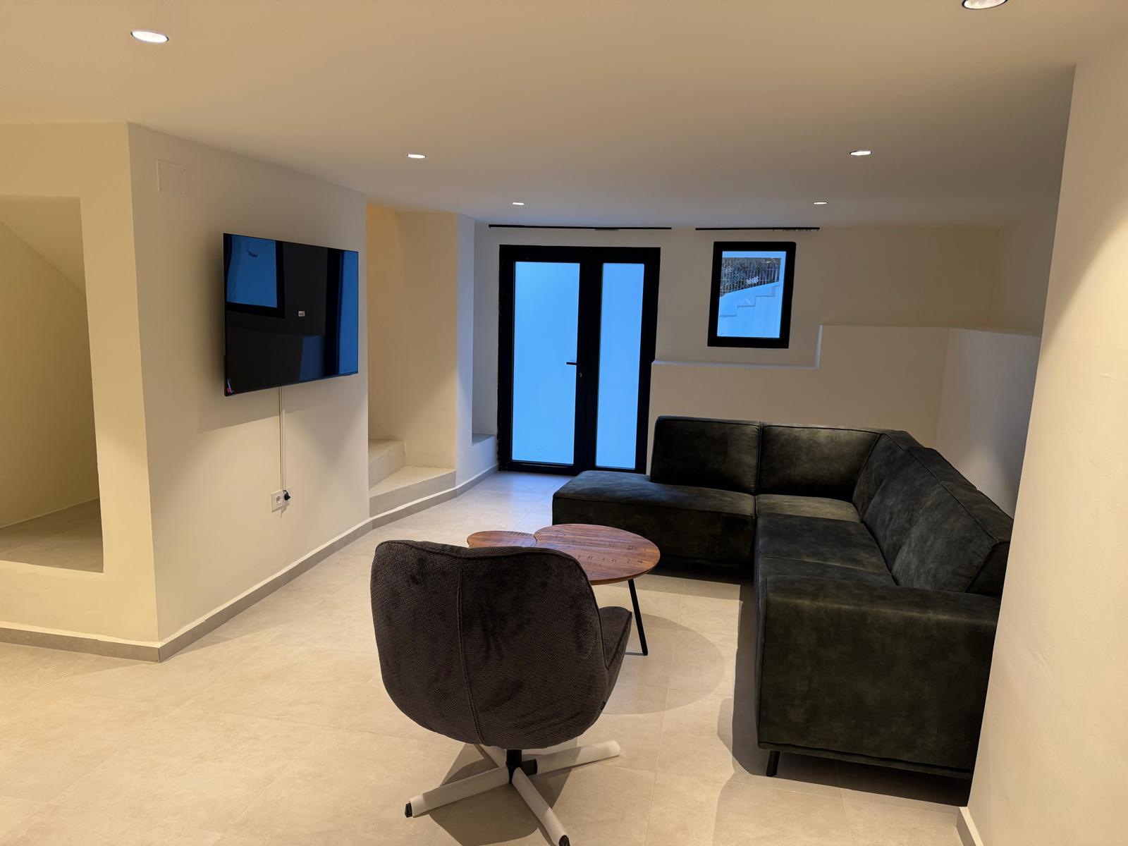 Modern lounge featuring a dark L-shaped sofa, swivel armchair, and wall-mounted TV. Recessed lighting and neutral tiling complement the minimalist interior design.