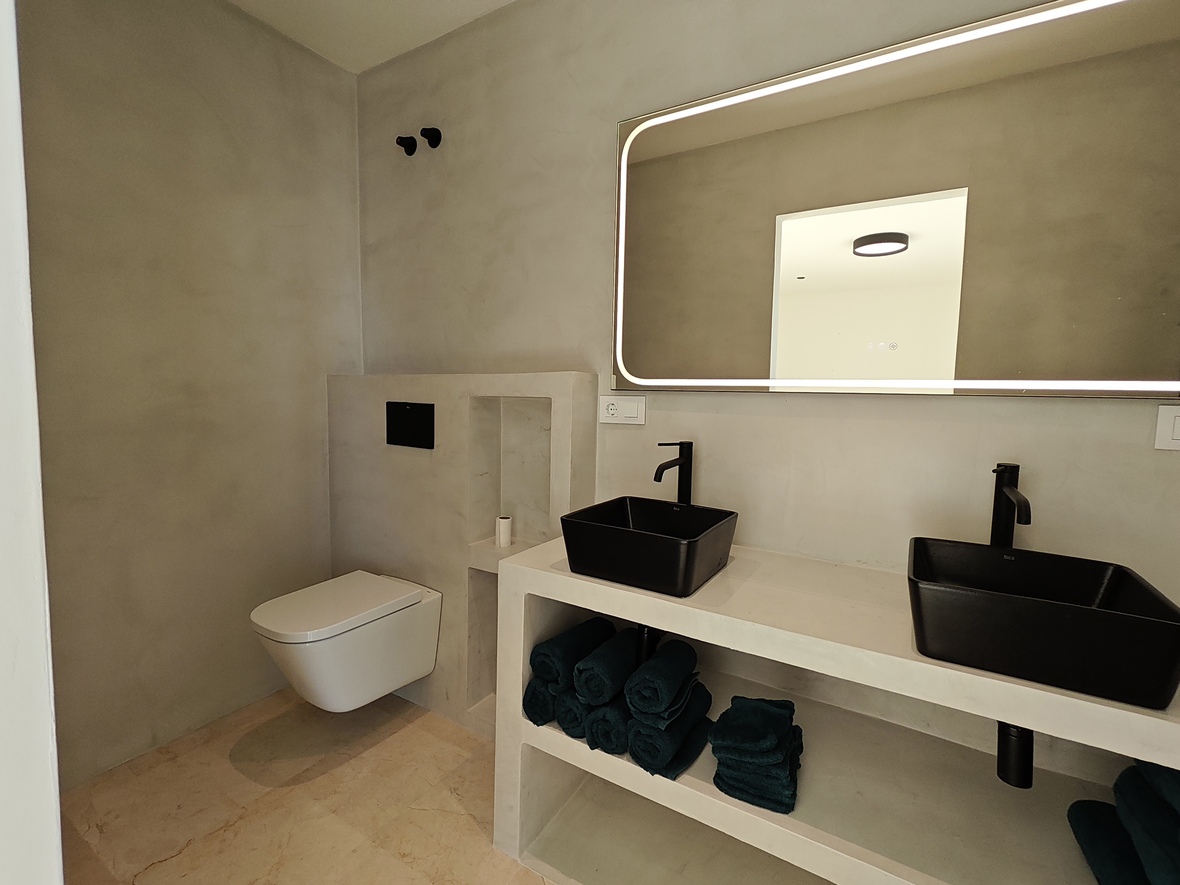Modern minimalist bathroom featuring grey microcement walls, double black vessel sinks on a built-in vanity, and a large backlit LED mirror.