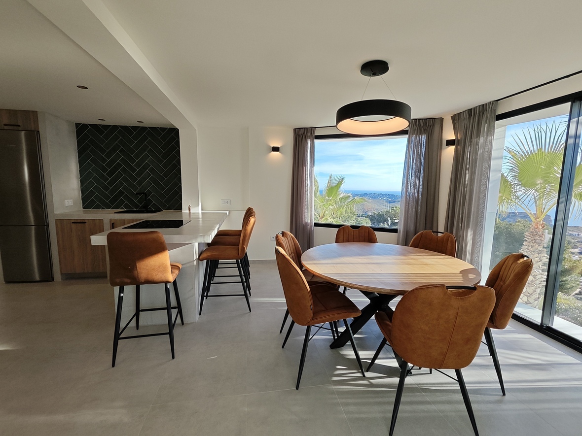 Modern open-plan dining area featuring a round wooden table, cognac chairs, and a designer kitchen with a dark herringbone backsplash and sea views.