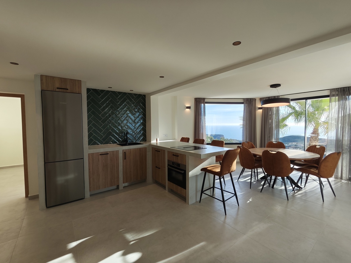 Modern open-plan kitchen and dining area featuring a green herringbone backsplash, integrated appliances, and a round wooden table with sea views.