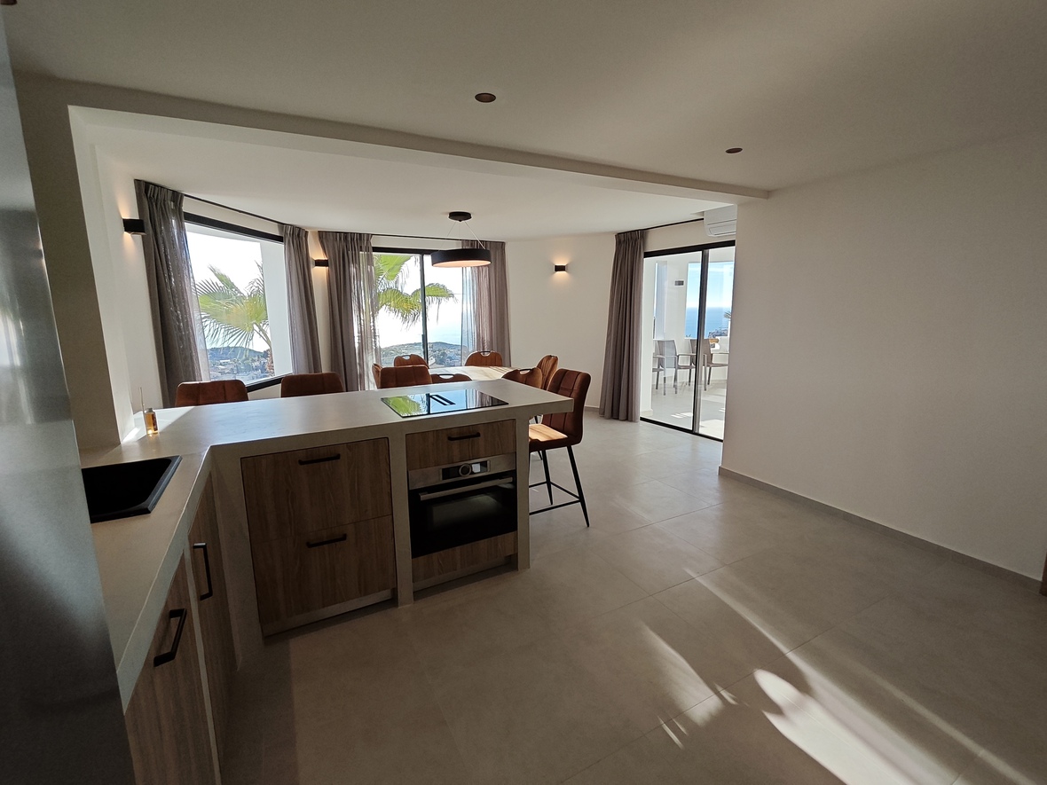 Modern open-plan kitchen with wooden cabinetry, integrated appliances, and a dining area offering panoramic sea views through large windows and a terrace door.