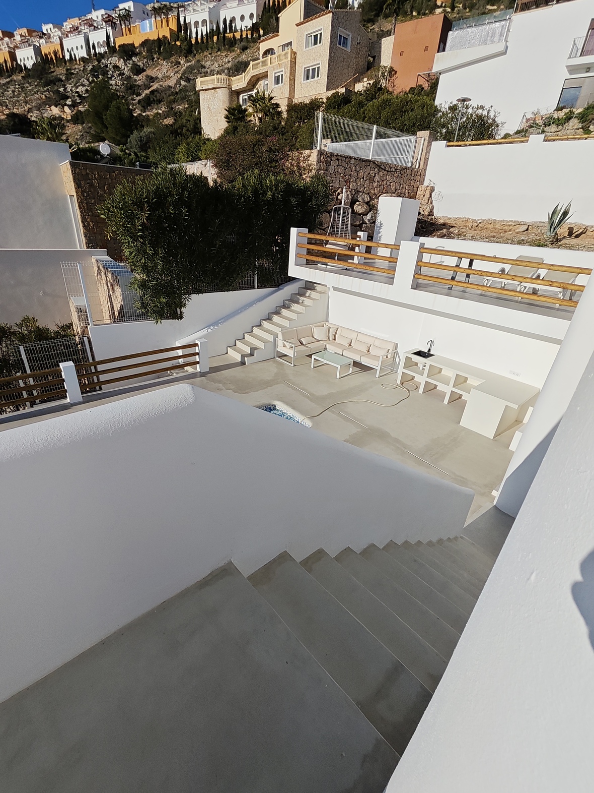 Multi-level outdoor terrace featuring white plaster walls, wooden railings, a lounge area with an L-shaped sofa, and a built-in outdoor kitchen under a hillside.