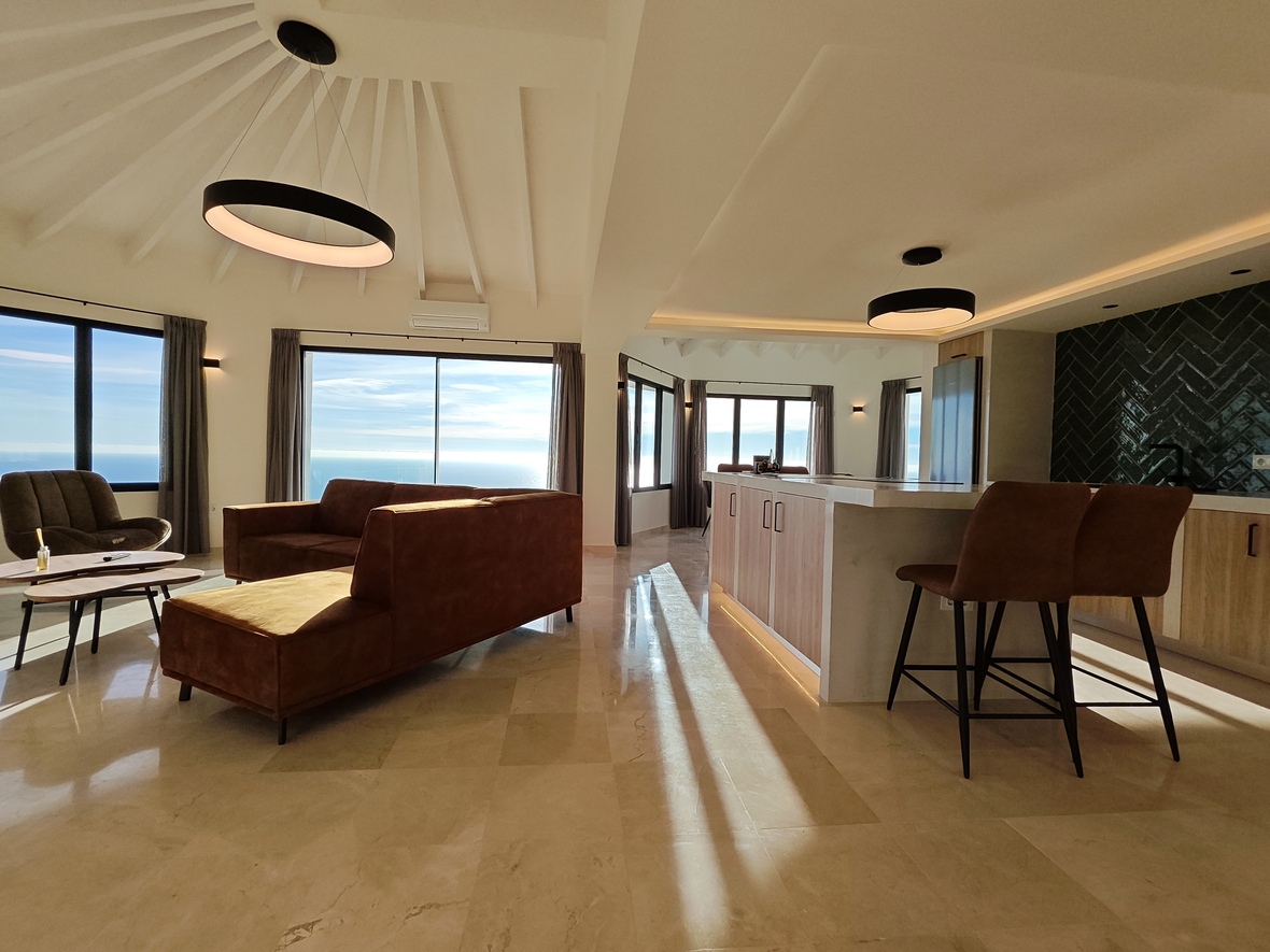 Open-concept living area featuring a vaulted beamed ceiling, marble kitchen island, and expansive floor-to-ceiling windows with panoramic sea views.