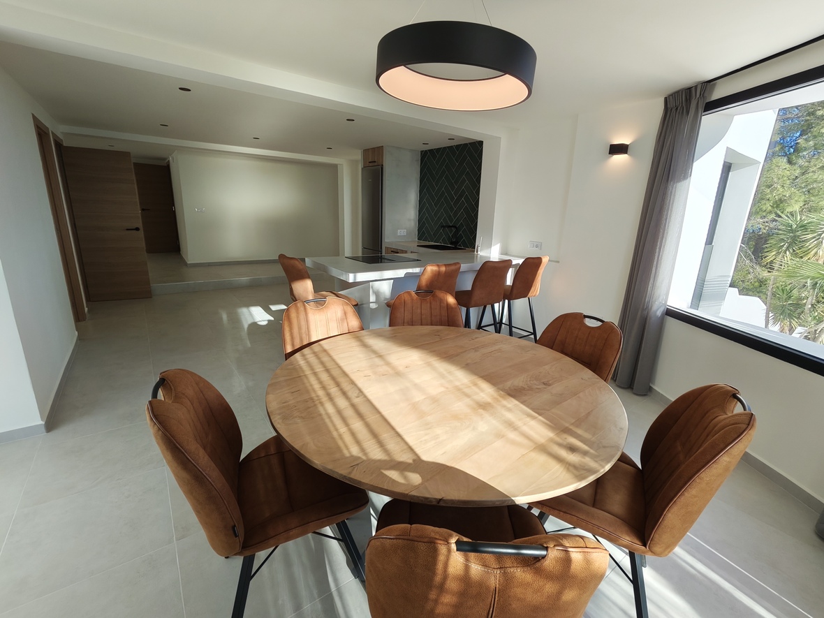 Open-plan dining area featuring a round wooden table, cognac-colored chairs, and a designer ring pendant, with a modern kitchen and breakfast bar in the background.