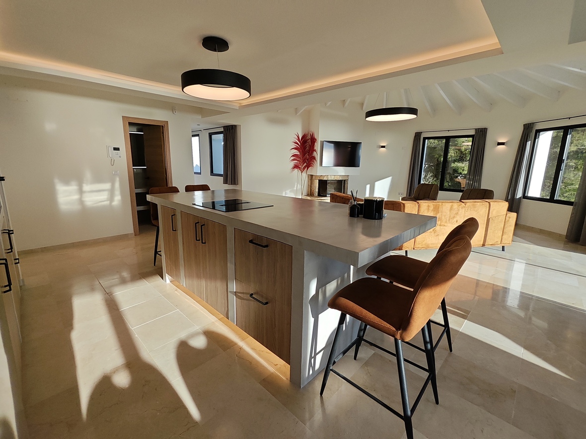 Open-plan kitchen with a concrete-finish island, integrated induction hob, and leather bar stools, set under a vaulted beamed ceiling with modern circular lighting.