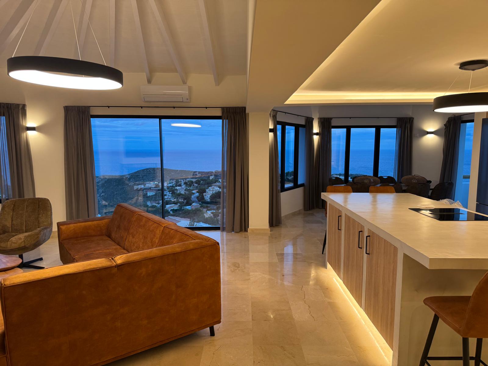 Open-plan living area featuring a cognac leather sofa and a kitchen island with integrated lighting, overlooking a coastal landscape through large windows.