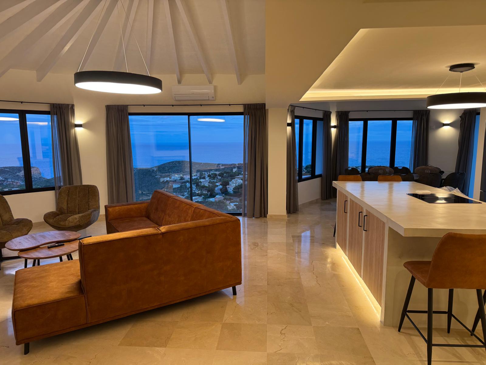 Open-plan living area featuring a cognac leather sofa, modern kitchen island with LED lighting, and panoramic sea views through large windows at dusk.