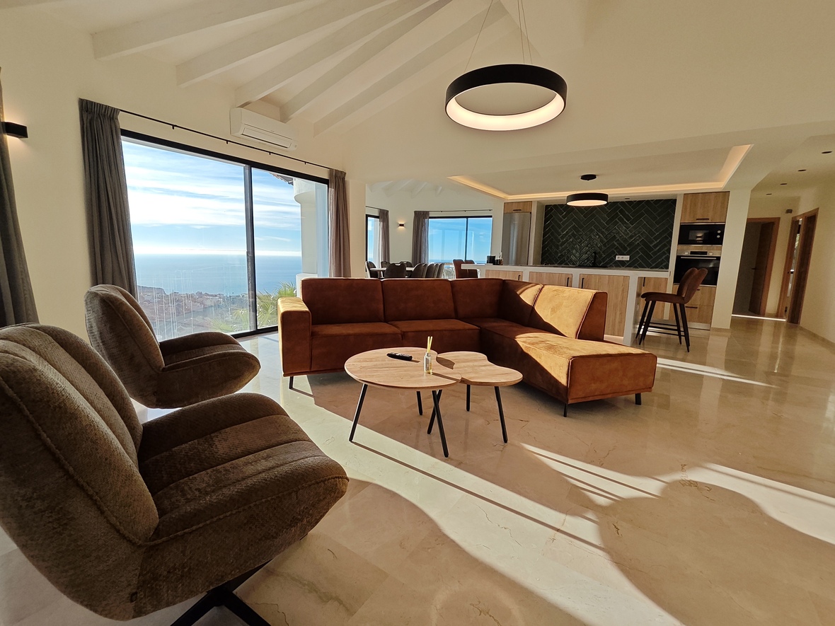 Open-plan living area with terracotta L-shaped sofa, vaulted beamed ceilings, and floor-to-ceiling windows offering sea views over polished stone floors.