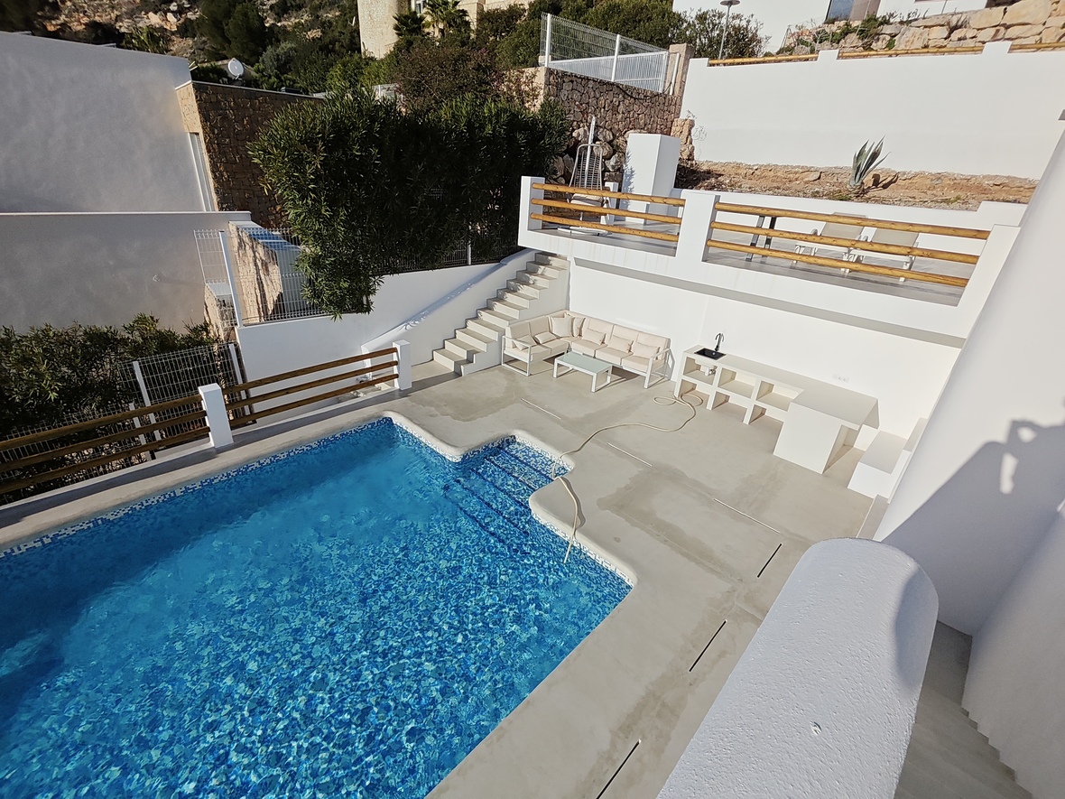 Outdoor pool area featuring a cement deck, integrated summer kitchen, and a tiered terrace with wooden railings and lounge seating.