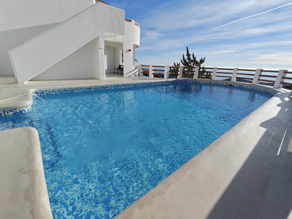 Outdoor swimming pool with blue mosaic tiling, featuring a white concrete terrace and a wooden railing overlooking the sea.