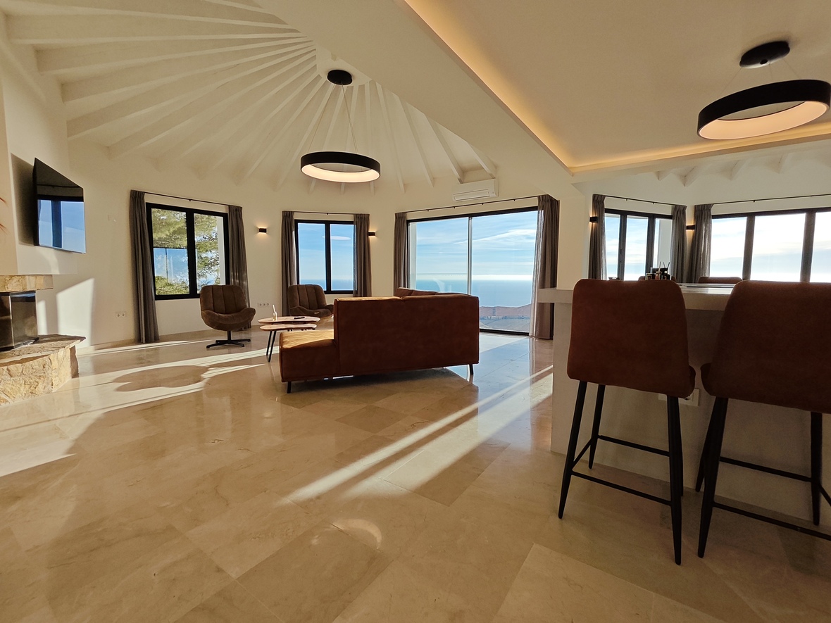 Spacious living area featuring a radial beamed ceiling, polished marble floors, and panoramic sea views. Includes a stone fireplace and modern circular lighting.