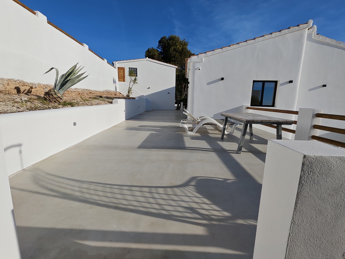 Spacious outdoor terrace featuring polished concrete flooring, white sun loungers, and a wooden dining table against a backdrop of Mediterranean white-washed architecture.