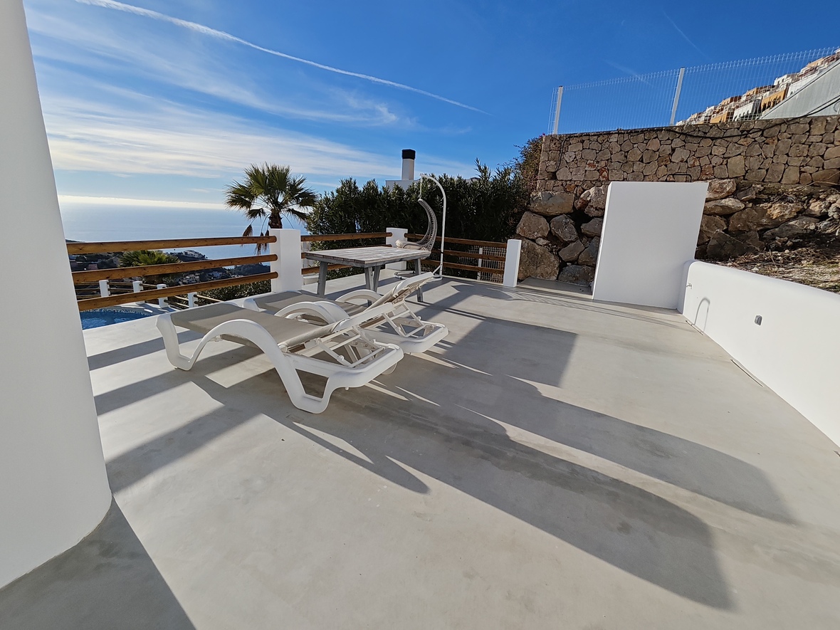Sun terrace featuring white loungers, a wooden dining table, and a hanging chair. Includes panoramic sea views, stone retaining walls, and smooth concrete flooring.