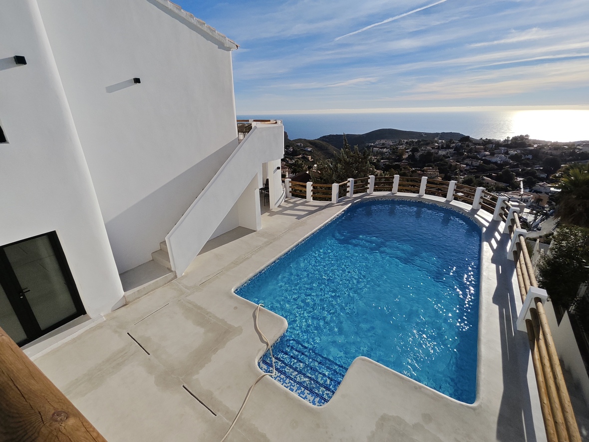 Swimming pool with blue mosaic tiles and a white concrete terrace, featuring wooden railings and panoramic views of the sea and coastal hills.