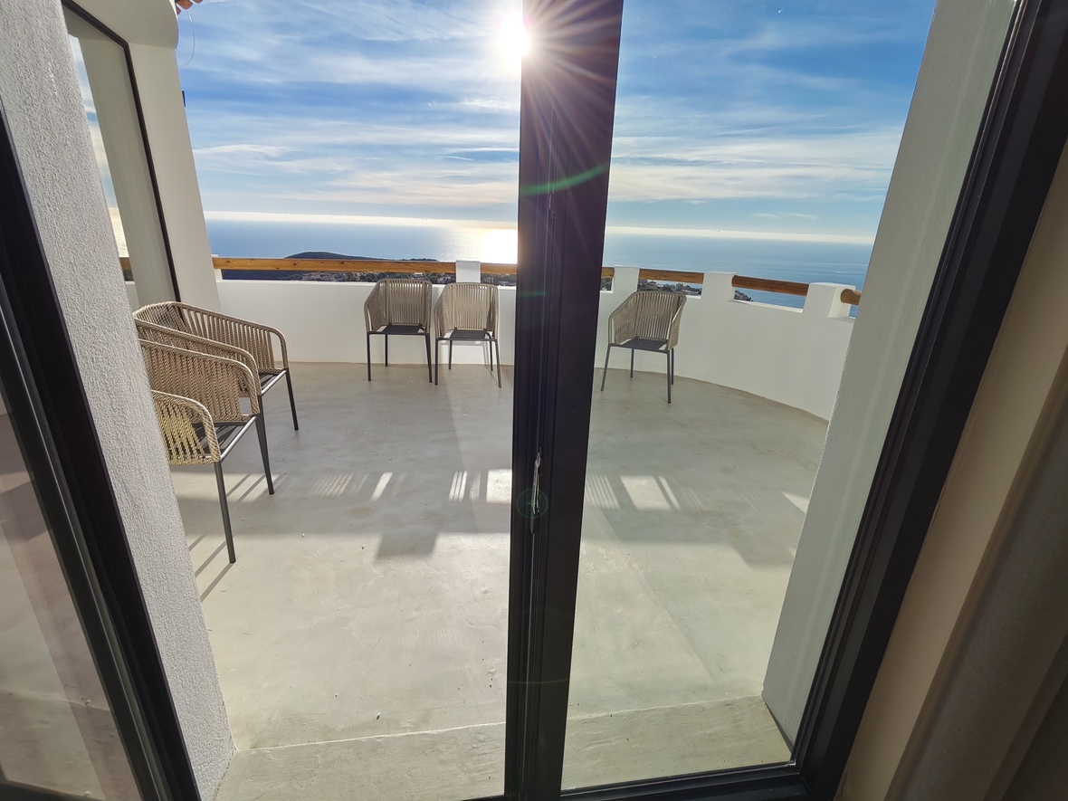 Terrace with panoramic ocean views, featuring woven outdoor chairs, white walls with wooden railings, and a polished concrete floor under direct sunlight.