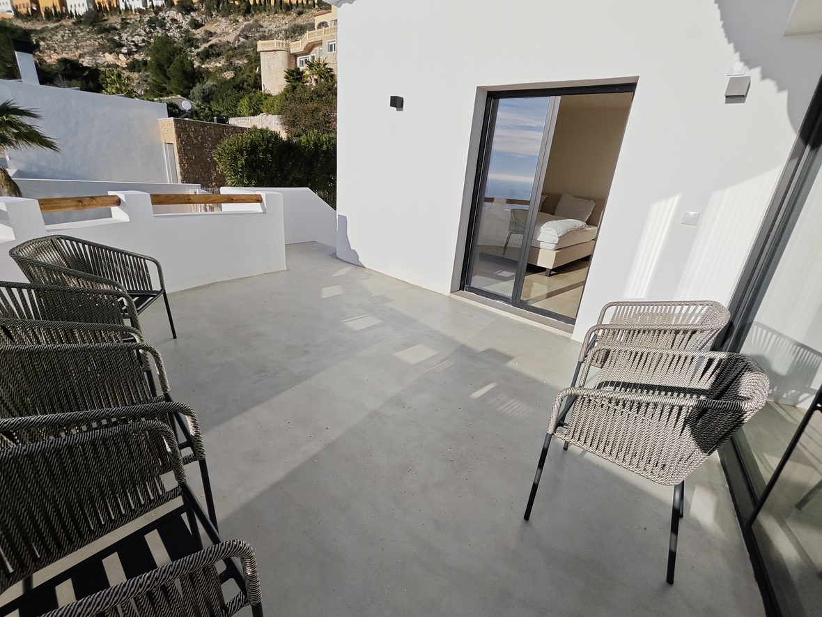 Terrace with smooth concrete flooring and modern woven chairs, featuring direct access to a bedroom via sliding glass doors against a white facade.