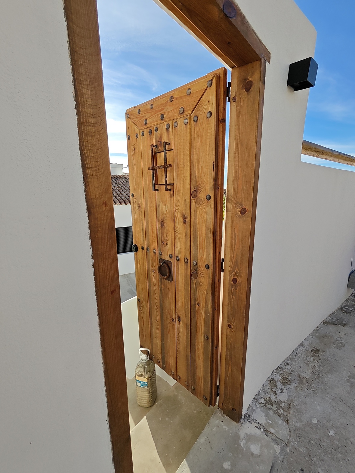 Traditional wooden door with iron studs and a small barred window, set in a minimalist white wall under a clear blue sky.