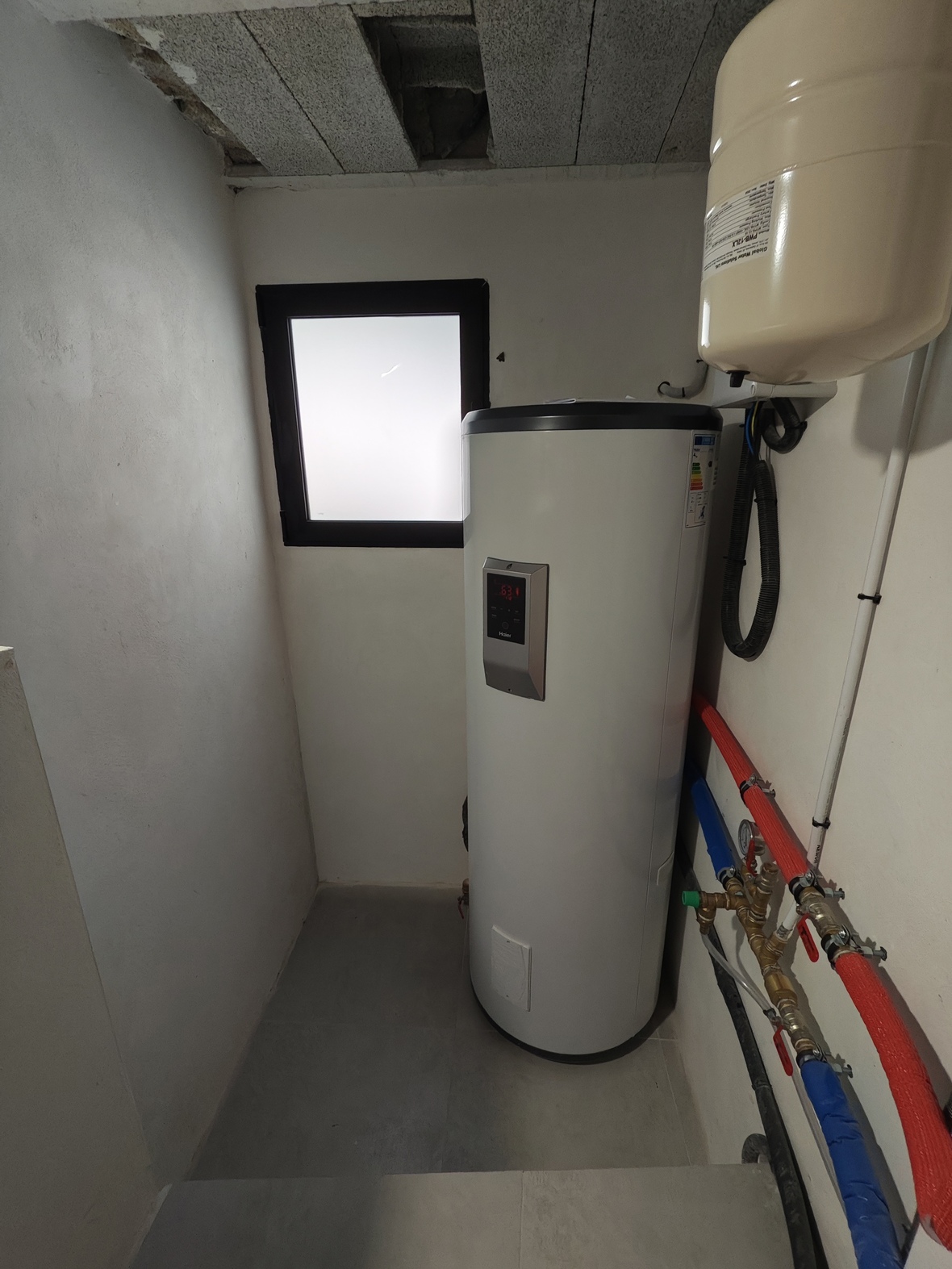 Utility room featuring a Haier aerothermal water heater, expansion tank, and insulated piping. The space includes a frosted window and grey tiled flooring.
