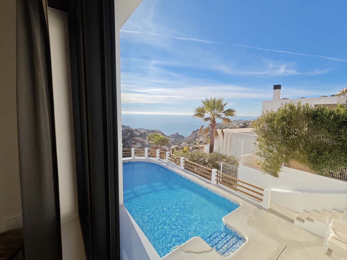 View from the interior across a turquoise swimming pool and white terrace, overlooking the Mediterranean coastline and palm trees under a clear sky.