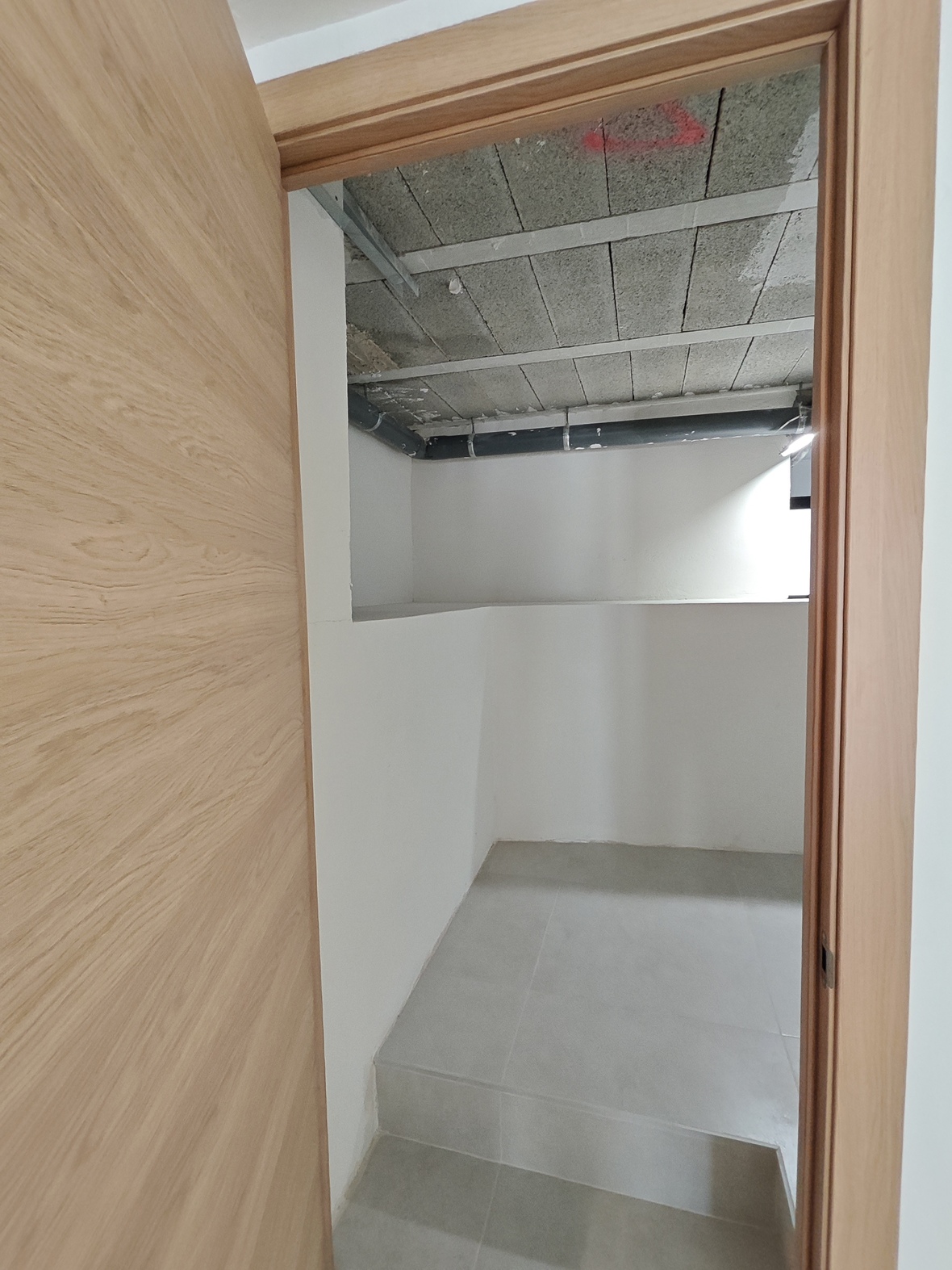 View through a light wood door into a utility or storage room featuring gray floor tiles, white walls, and exposed ceiling infrastructure with plumbing.
