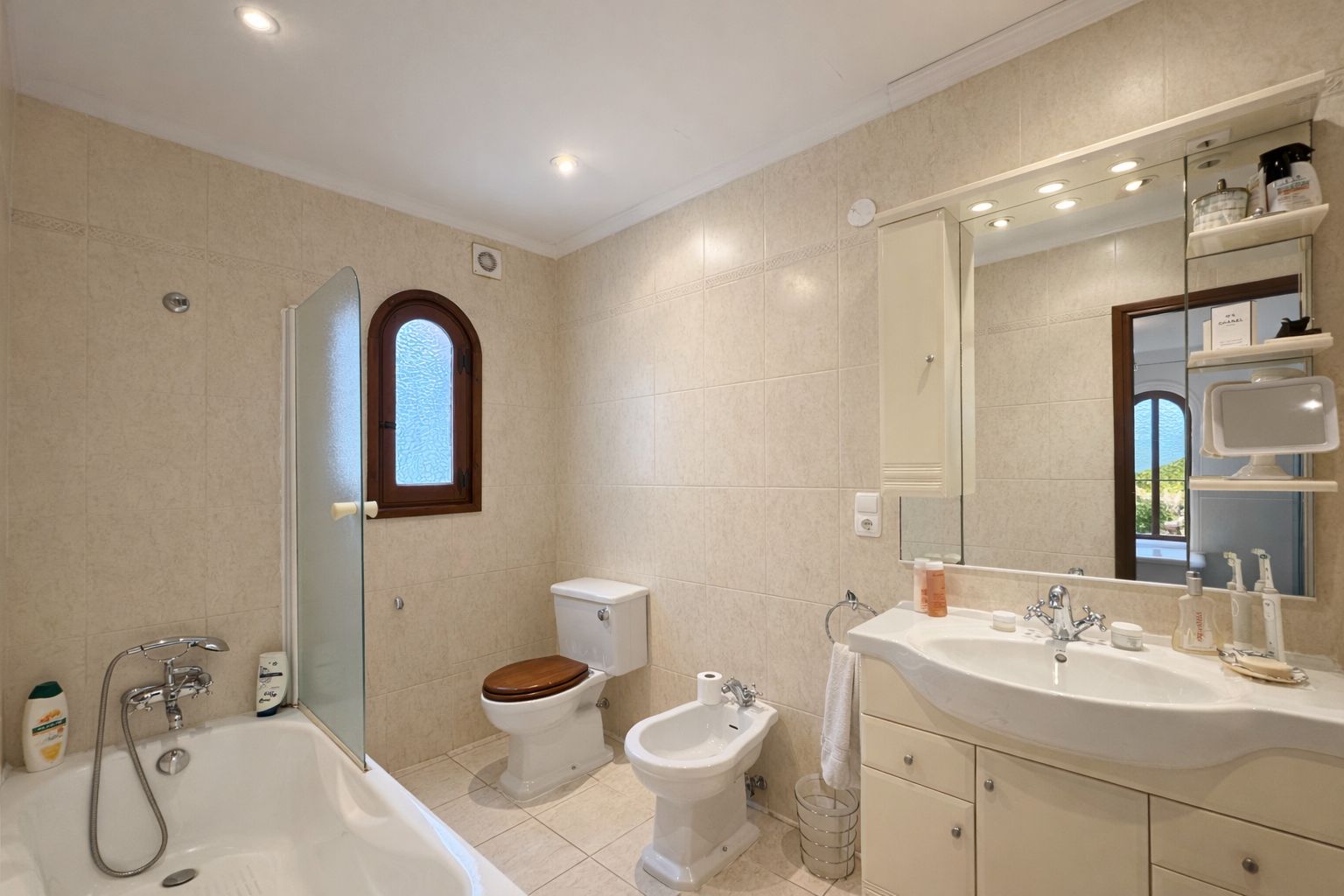 Bathroom featuring a bathtub with glass screen, bidet, and toilet with wooden lid. Includes a vanity with integrated sink, large mirror, and an arched wooden window.