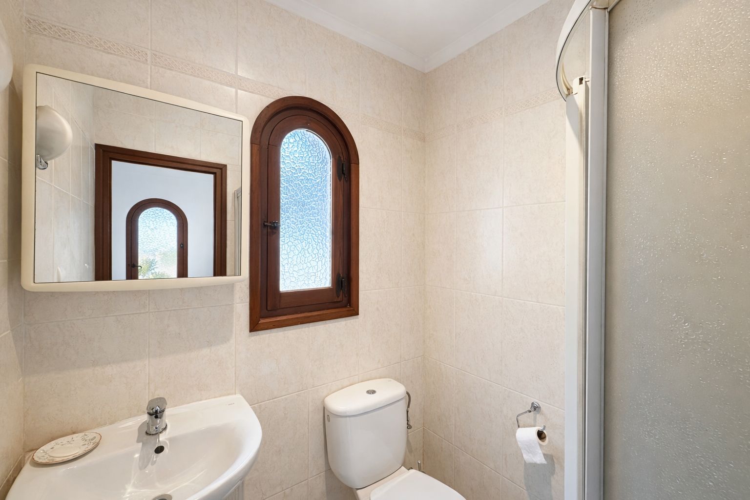 Bathroom featuring cream-toned wall tiles, a wooden arched window with textured glass, and a white ceramic pedestal sink.