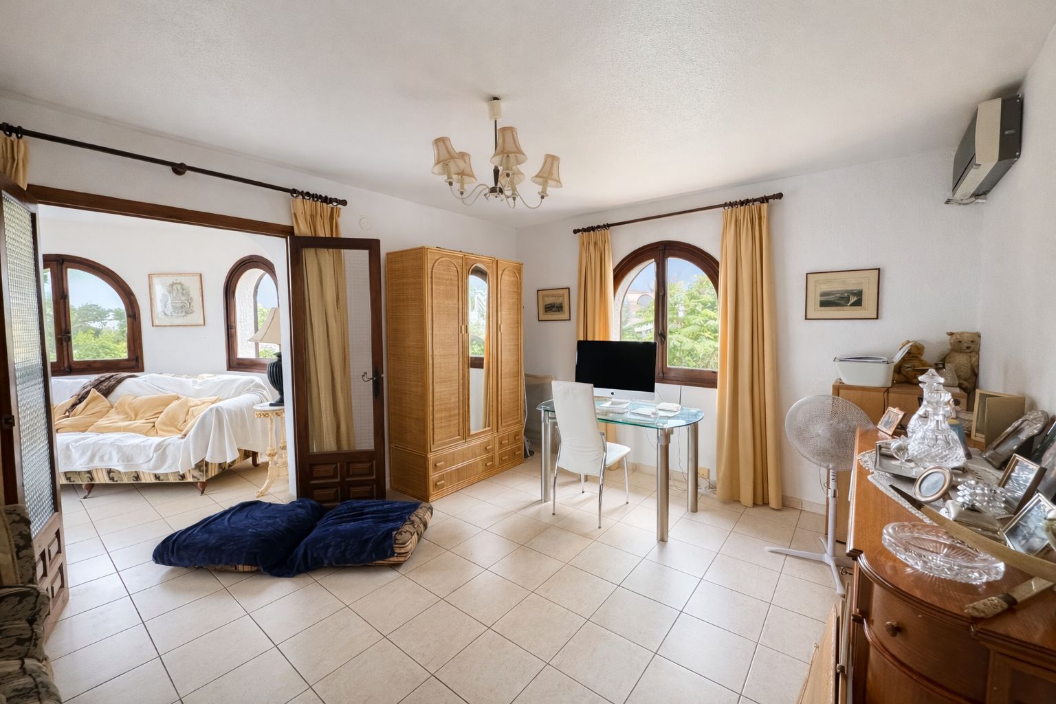 Bright home office featuring a glass desk, wooden wardrobe, and arched windows. Includes tiled flooring and direct access to an adjacent lounge area.