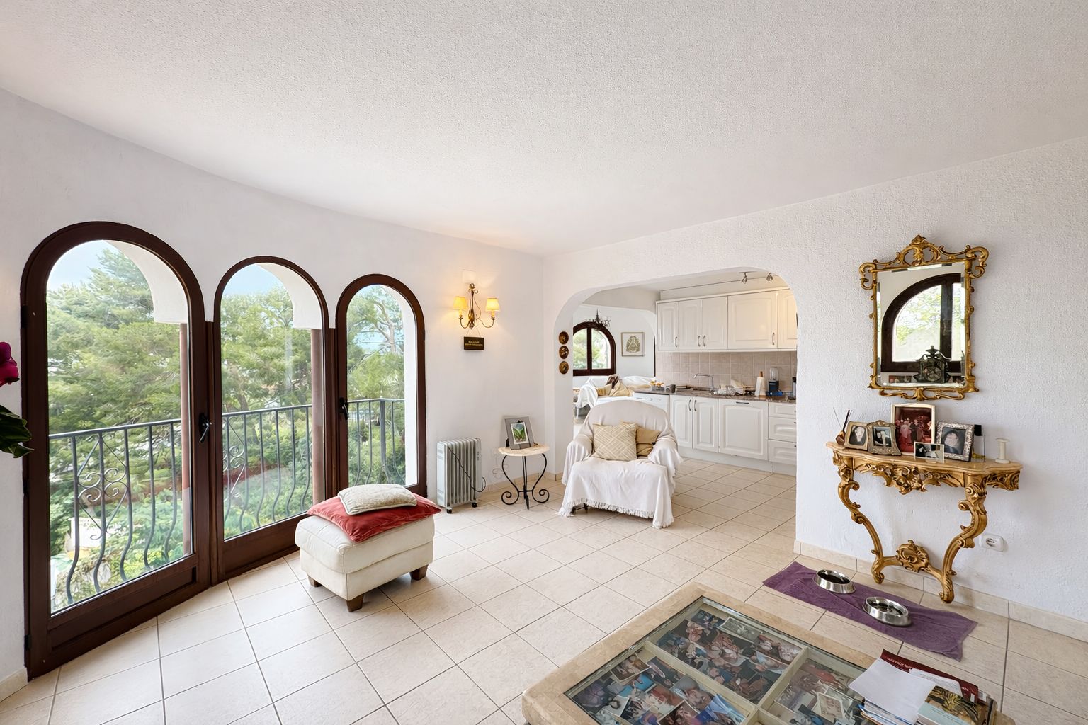 Bright living area featuring three arched glass doors opening to a balcony with garden views, white textured walls, and an ornate gold console table.