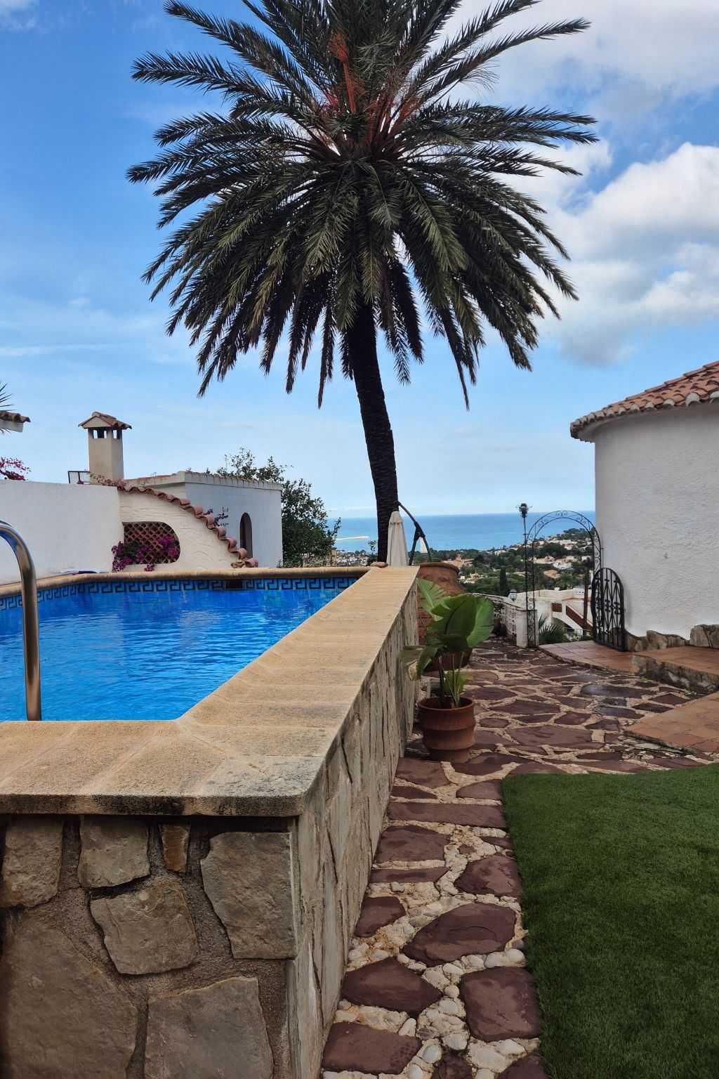 Elevated swimming pool with stone cladding and blue tiling, featuring a flagstone terrace, a large palm tree, and views of the Mediterranean coastline.