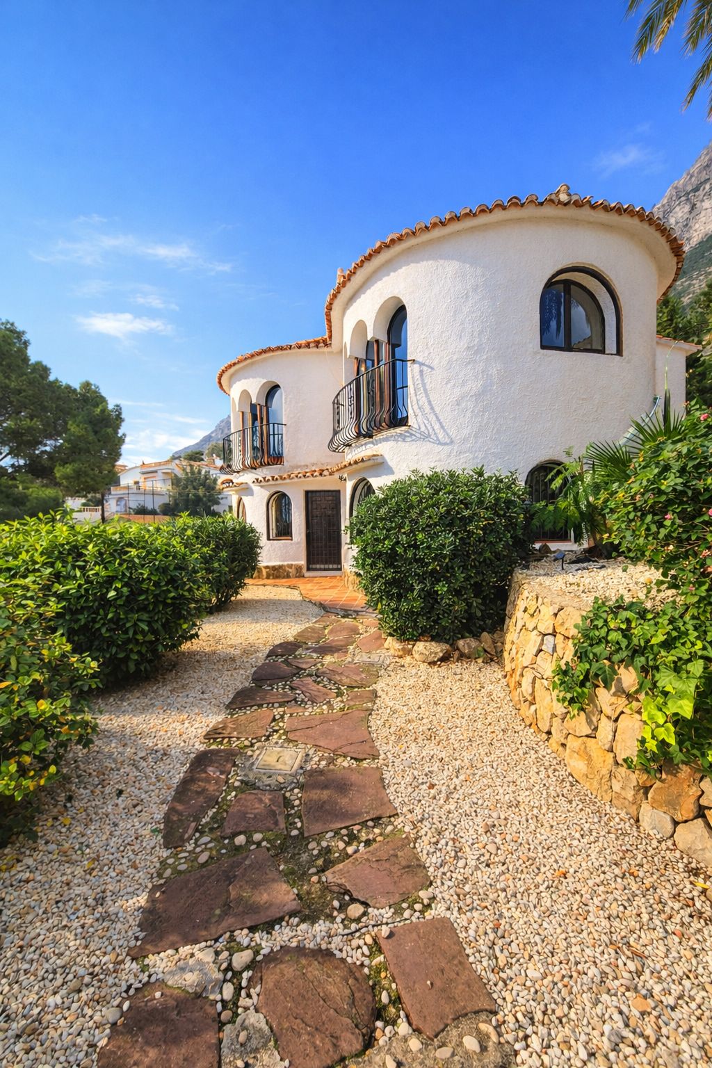 Exterior view of a Mediterranean villa featuring curved white walls, arched windows, and wrought iron balconies. A flagstone path leads through gravel landscaping.