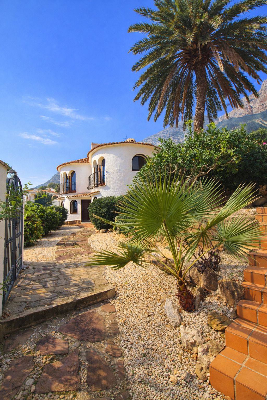 Exterior view of a white Mediterranean villa with rounded towers, arched windows, and a stone pathway set within a xeriscaped garden with palm trees and mountain views.
