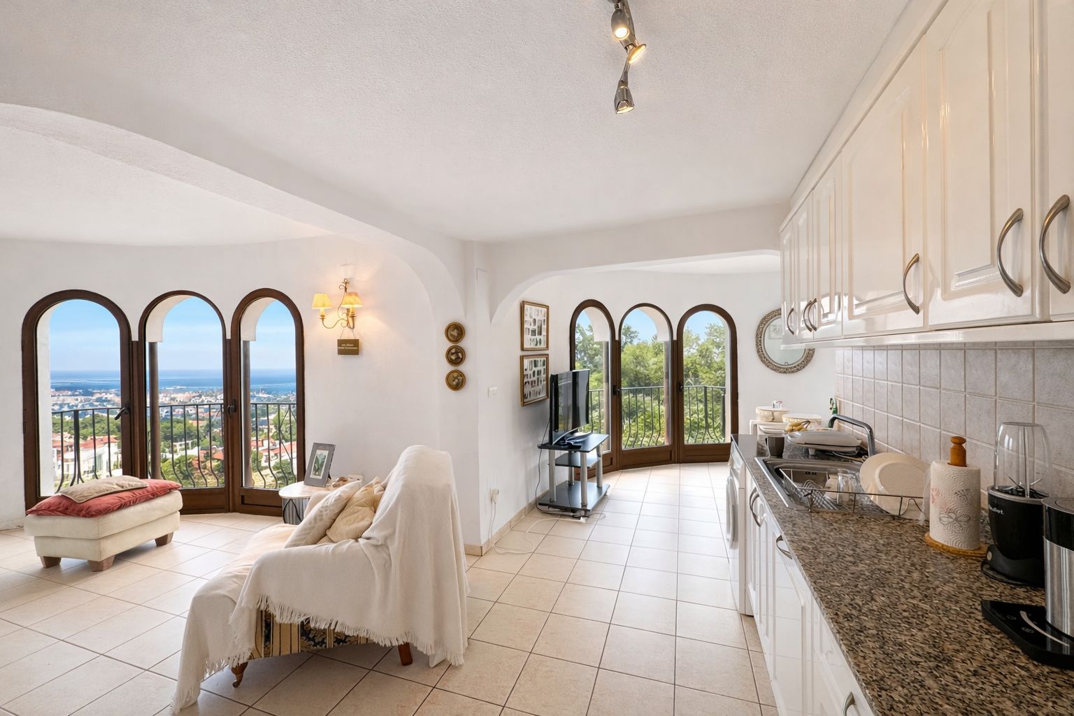 Open-plan living area featuring a kitchen with granite countertops and white cabinetry, alongside arched windows offering panoramic sea and coastal views.