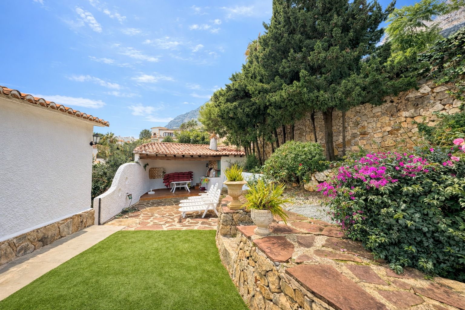 Outdoor terrace featuring natural stone paving, artificial turf, and a covered porch with terracotta tiles. Stone walls and Mediterranean flora frame the space.