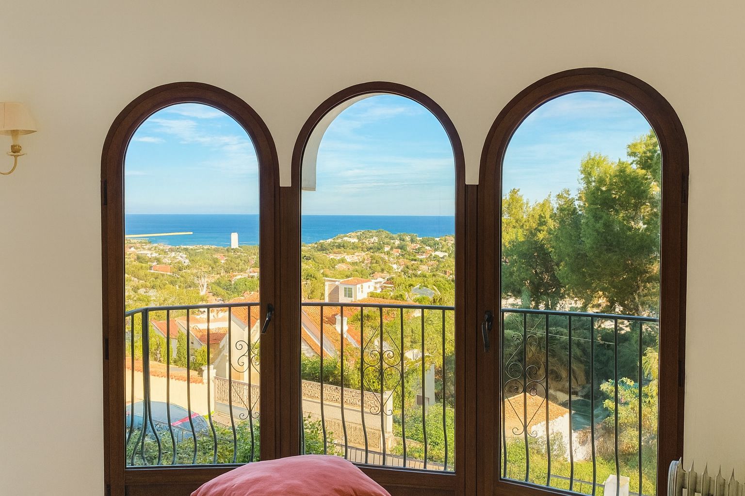 Three arched windows with dark wood frames provide an elevated view of the Mediterranean coastline, featuring a wrought iron railing and lush green surroundings.
