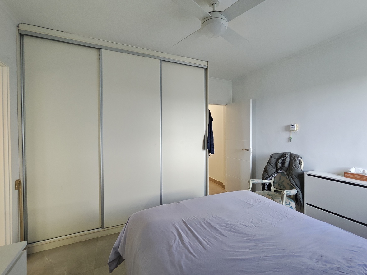 Bedroom featuring a large three-door sliding wardrobe, a white ceiling fan, and a classic armchair positioned beside a minimalist chest of drawers.